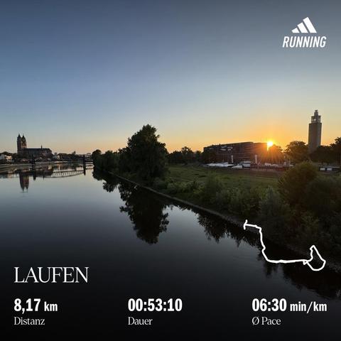 Am Horizont eine Kirche (Magdeburger Dom), im Vordergrund, ein nahezu spiegelglatter Fluss (die Elbe) darüber die aufgehende Sonne gleich rechts hinter einem großen Gebäude (Stadthalle und Baustelle), der Fluss ist gesäumt von grünen Bäumen, Büschen und Wiesen, die Stadtsilhouette von Magdeburg über die Elbe Richtung Dom geblickt am frühen Morgen