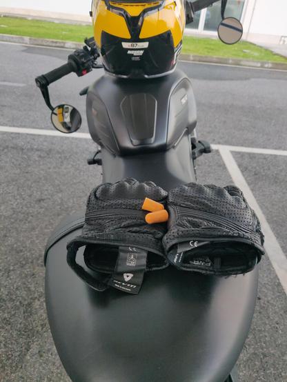 The image shows a black motorcycle with a yellow and black helmet placed on the handlebars. The motorcycle is parked on a paved surface with a grassy area and a building visible in the background. On the seat of the motorcycle, there is a pair of black motorcycle gloves with perforations, and two orange earplugs are placed between the gloves. The gloves have a tag with the brand name "REV'IT!" and a CE mark, indicating they meet European safety standards. The motorcycle's seat is black and appears to be made of a smooth, matte material. The handlebars have a round mirror on the left side, and the motorcycle's body has a sleek, modern design.