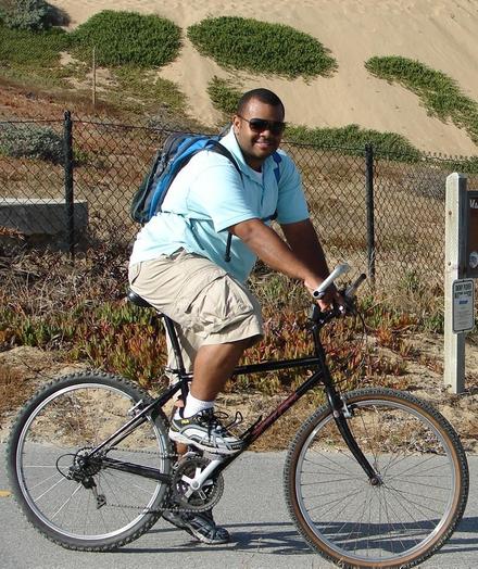 A man smiles as he pauses on his bike ride to greet us. He is on the Sand City section of the Monterey Bay coastal trail, and a tall sand dune is behind him. He is wearing dark sunglasses, a light blue short-sleeved shirt, beige pedal-pusher length shorts, white socks, white-silver-black tennis shoes, and a bright blue day pack. His skin is black.