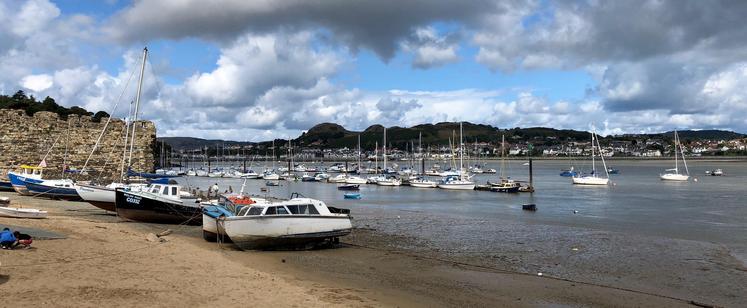 This picture showcases a serene coastal scene featuring a sandy beach in the foreground with several boats grounded on the shore, likely due to low tide. Among the boats, names like "WELSH MAID" and "Pandora" are visible. To the left, there's an old stone wall, possibly part of a historic fortification or castle. The water is calm, dotted with numerous moored sailboats and small vessels. Across the water, a quaint town with white buildings nestles against green hills, creating a picturesque backdrop under a partly cloudy sky, with patches of blue peeking through.