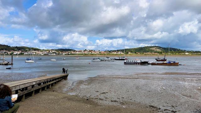 This picture captures a serene coastal scene in Gwynedd, Conwy, Wales. The image showcases a wide estuary during low tide, with several small boats and sailboats anchored in the shallow waters. A wooden jetty extends into the water from the lower left, where a couple of people are standing, enjoying the view. In the foreground, a person is seated on the left, partially visible, holding a white container, possibly with food.

Across the water, a charming town with rows of white houses and red-roofed buildings sits at the base of gently rolling green hills. The sky above is dramatic, filled with a mixture of thick, fluffy clouds and patches of blue, casting dynamic lighting over the landscape. The overall atmosphere is peaceful and picturesque, typical of coastal Welsh scenery.