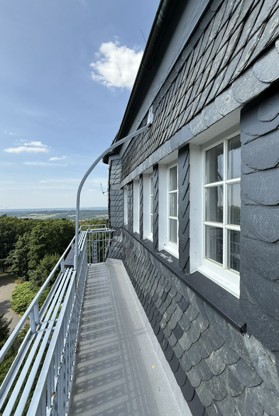 Das Bild zeigt eine seitliche Ansicht des Danzturms in Iserlohn mit einem weiten Blick über die Landschaft. Der Turm ist mit Schiefer verkleidet und hat weiße Fensterrahmen. Ein Balkon mit Metallgeländer erstreckt sich entlang der Seite, von dem aus man die grüne, bewaldete Umgebung überblicken kann. Der Himmel ist klar mit einigen Wolken, was die Aussicht noch beeindruckender macht.