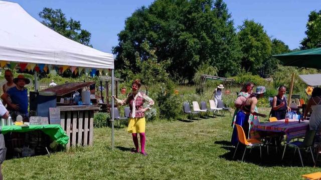 Une clown au milieu d'un jardin fleuri, quelques minutes avant son spectacle.
Elle est habillée d'un short jaune, collants framboise, un gilet à carreaux rouge, un chapeau rond, un chemisier et un nez rouge
Des stands festifs à droite et à gauche, plusieurs personnes à chaque stand, des grands arbres en fond et du ciel parfaitement bleu