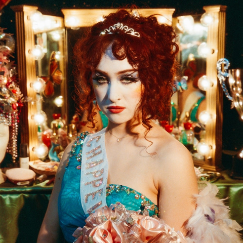 “The Rise And Fall Of A Midwest Princess” by Chappell Roan. It's Chappell sitting in front of a dressing table with several round light bulbs around the mirror. She's wearing white foundation, very glittery eye shadow, rouge and red lipstick and has a small tiara in her hair. Her dress is turquoise and she's wearing a sash with her name in glitter letters.