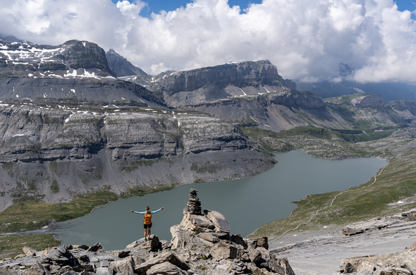woman standing on a rock and spreading her arms, below a mountain lake and high mountains in the background. some clouds above.