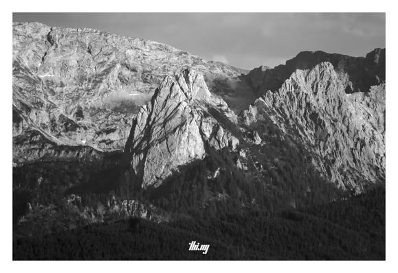 B&W photo of a large mountain massif (Hochplatte, 2079m) rising up behind lower ridges of dark dense forest. Inbetween, the imposing tooth-like Geiselstein rock.