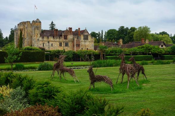A picturesque view of Hever Castle in Kent, England, on a slightly overcast day. The historic stone castle, with its flag flying, stands prominently in the background amidst lush green trees. In the foreground, a vibrant green lawn features four rustic, life-sized deer sculptures crafted from woven branches. A pond or water feature is visible between the lawn and the castle, surrounded by neatly trimmed hedges and diverse foliage. The scene evokes a sense of tranquil, historic grandeur.
