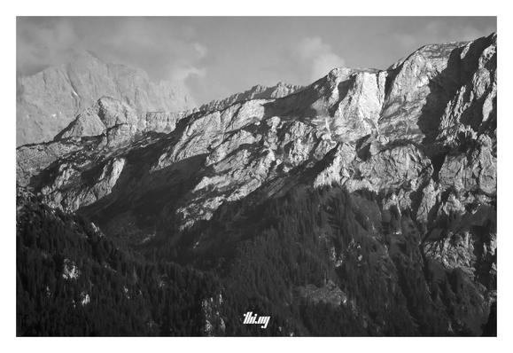 B&W photo of the north-eastern side of Hochplatte with the forested valley in front, and the Zugspitze (Germany's tallest mountain) in the hazy background, its peak surrounded by clouds.