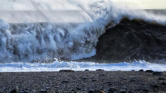 A dramatic close-up of a massive, dark wave crashing and breaking towards a stony beach. The wave's crest is white with foam and spray being blown back by the wind, illuminated by sunlight from the upper right, while the dark body of the wave shows its immense power. The foreground features a pebbled beach stretching across the bottom of the frame.