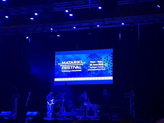 A concert stage is being set up in blue light. On the screen shows Matariki Festival, 20 June 2025 and the venue.