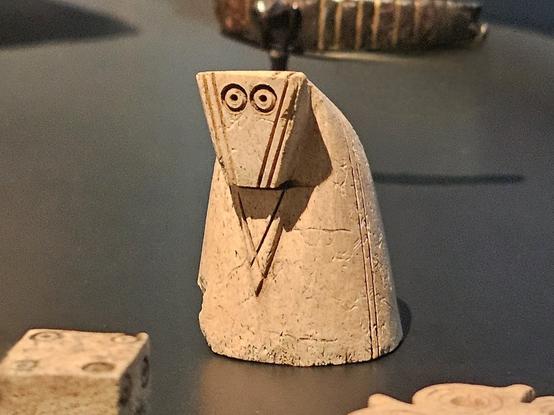 A carved chess piece representing a knight, depicted in an abstract, stylized form with a triangular head, wide circular eyes, and incised lines suggesting folded arms. The figure has a hunched, cylindrical body and is displayed on a dark surface with other game pieces nearby,