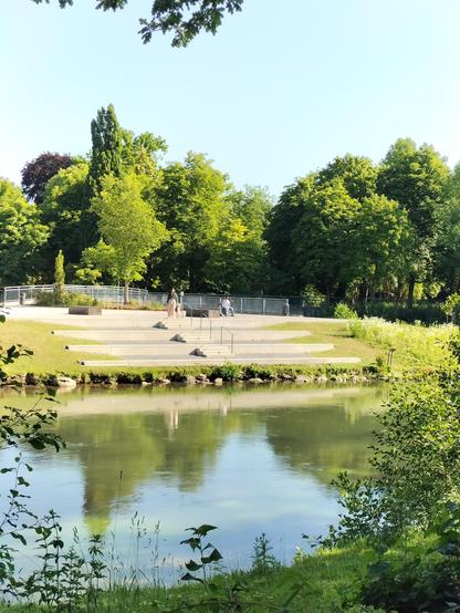 Im Vordergrund ein langsam fließendes Gewässer. Dahinter ein mit Steinen befestigt es mit Gras und niedrigen Pflanzen bewachsenes Ufer, dann drei große sitzhohe Betonstufen, die mit Treppen und einem Geländer verbunden sind. Im Hintergrund hohe Bäume.