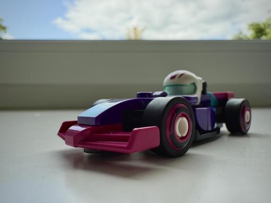 Alt Text von ChatGPT:

Das Bild zeigt ein kleines Spielzeugauto im Stil eines Formel-1-Rennwagens. Das Auto besteht aus bunten Bausteinen aus LEGO. Die Farben sind dominierend lila, pink und blau, mit schwarzen Reifen und weißen Felgen. Im Cockpit sitzt eine kleine Figur mit einem weißen Helm, der ein Visier in Türkis hat. Der Hintergrund zeigt ein Fenster mit teilweise bewölktem Himmel und etwas Grün von Bäumen. Das Licht fällt von draußen herein, was für einen interessanten Kontrast zwischen dem hellen Hintergrund und dem schattigeren Vordergrund sorgt.