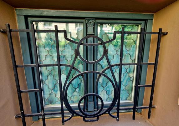 Fenster von innen, mit Jugendstilelementen. Dabei zeigt die Scheibe ein grünlich-türkisfarbenes Muster. Vor der Scheibe ist eine metallene Vergitterung.