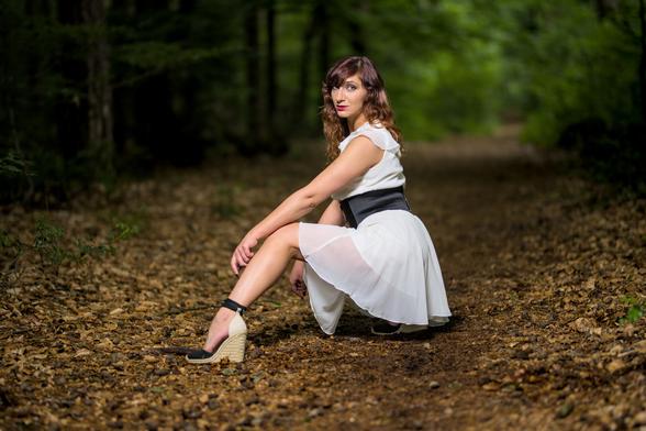 Woman in a white dressis kneeing with front leg extended on a path in a forest.