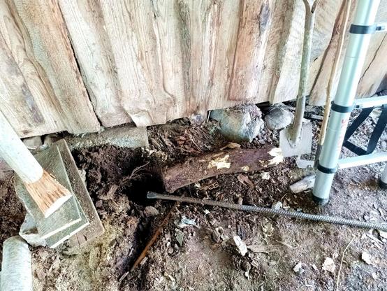 An old log that has been extracted from half a meter deep in the ground. It's rotten and lost about half its diameter, but this one still held. There's a temporary support on the left with a wedge and some planks propping up a wooden post. A ladder stands on the right. A hole and some leverage lay next to the rotten stump.