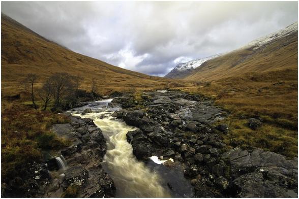 A scenic view of a winding river flowing through rocky terrain, surrounded by rolling hills covered in golden grasses and patches of snow. The sky is overcast with dark clouds.