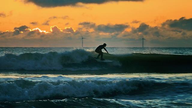 A silhouette of a surfer rides a wave at sunrise. The sky is a dramatic orange and gold with dark clouds, and two wind turbines are visible in the distance on the horizon. The ocean is a mix of dark blues and whites from the breaking waves.