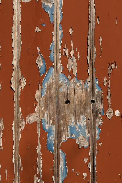 Wooden planks on the side of a shed, the brown paint is peeling off to reveal blue underneath and the wood below that. In the middle of an almost circular bare patch are two round holes that resemble eyes