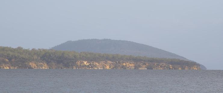 A wide landscape view over calm waters toward distant tree lined hills.  The closer headland's low sandstone cliffs are lit by the late afternoon sun, giving them a golden glow, extending to the trees above.  A taller headland rises behind, hazy in the distance and shadowed by cloud out of frame.