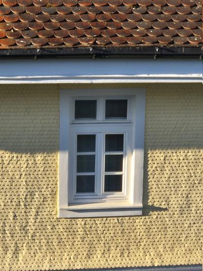 In einer Hausfassade aus gelb gestrichenen Holzschindeln befindet sich ein einzelnes weißes, doppelflügliges Sprossenfenster. Über dem Fenster befindet sich ein mit roten Biberschwanzziegeln eingedecktes Dach.