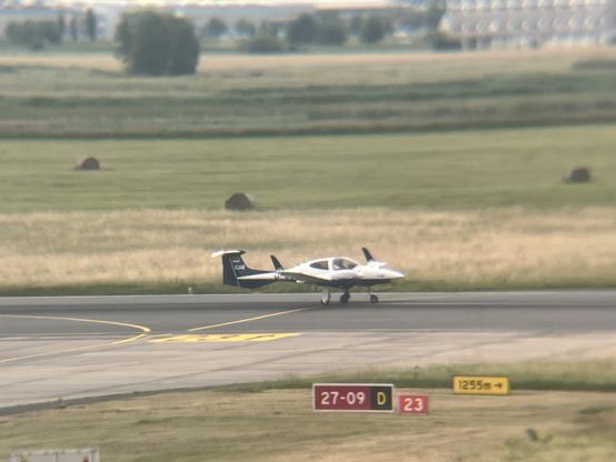 A small aircraft is taxiing on a runway, with grassy fields in the background. The plane has a white and dark blue design and features visible registration numbers. Nearby signage indicates runway information.
