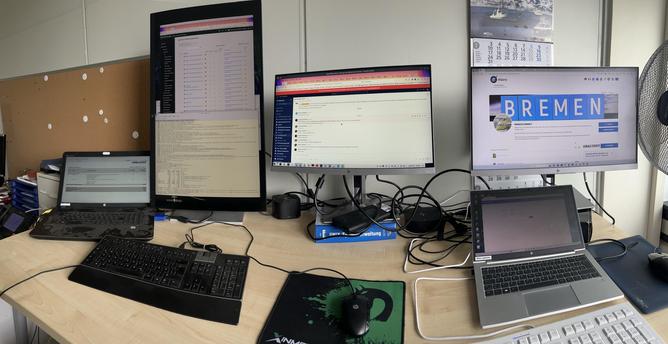A workspace featuring multiple computer monitors and laptops. One large monitor displays a coding interface, another shows a chat application, and a third presents a community page for Bremen. The desk is cluttered with cables, a keyboard, and a mouse. A calendar