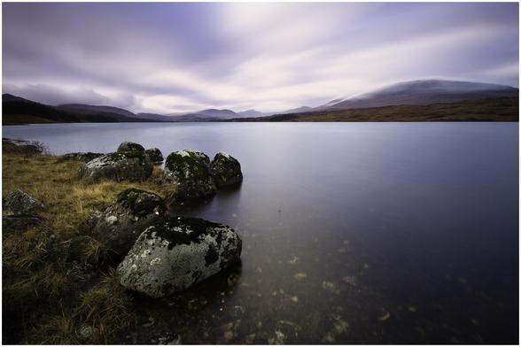 A serene landscape featuring a calm loch bordered by moss-covered rocks and dry grass. In the background, rolling hills and mountains are shrouded in soft clouds, creating a tranquil atmosphere. The sky is partly cloudy, with gentle hues.