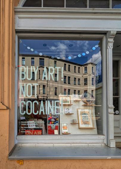 A quirky shop window reflecting a cloudy sky and the facade of a building opposite. Large white text on the window reads "BUY ART! NOT COCAINE!". Inside, various artworks and items are displayed on shelves and hanging. A sign in the bottom left, partially obscured, seems to be in German, potentially advertising the shop's offerings, while another small sign says "No magic!". The reflection of blue sky and clouds is visible above the text, and a string of colourful circular decorations is draped across the top of the window. The building's ornate architectural details frame the shopfront.