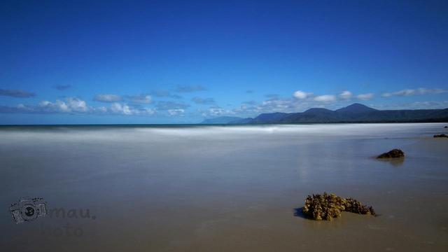 Sandy beach with a calm sea under a clear blue sky. Mountains are visible on the horizon, and small patches of seaweed are scattered across the wet sand in the foreground. Sparse clouds dot the sky.