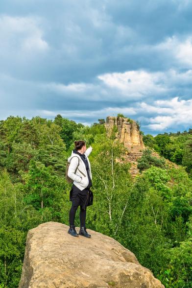Eine weibliche Person im Vordergrund auf einer Klippe zeigt mir dem Finger auf eine Sandstein Klippe im Hintergrund. Dazwischen geüner Wald.