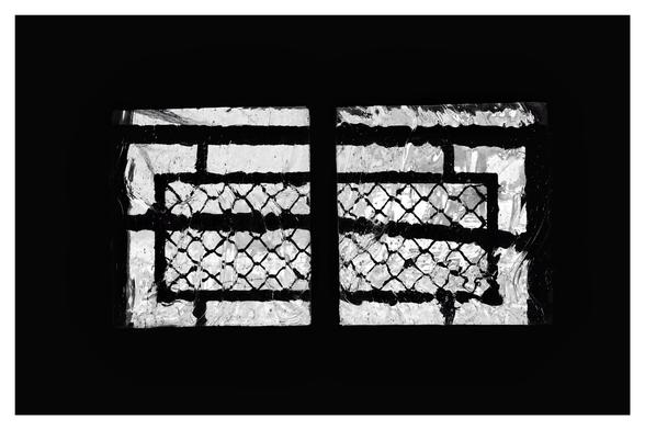 Schwarz-Weiß-Foto im Querformat mit weißem Rand. Zu sehen ist ein querformatiges Fenster mit strukturiertem Glas und einem schwarzen Gitter davor. Die Wand um das Fenster beim ist schwarz.