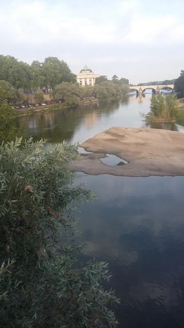 Photo d'une grosse rivière avec une étendue sableuse au milieu qui forme une île.
Un bâtiment et un pont qui ont l'air anciens sont en arrière-plan. À gauche, la berge, qui semble être un parking