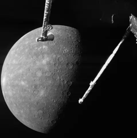 Eine etwas über zur Hälfte beleuchtete Merkurscheibe. Die beleuchtete Seite zeigt zum linken Bildrand. Die Scheibe des Planeten befindet sich am unteren linken Bildrand. Die Oberfläche ist fest, stark verkratert, und in hellen Grautönen. Aus der Schwärze des Weltraums darum herum ragen von rechts oben nach schräg unten zwei Arme einer Raumsonde ins Bild.