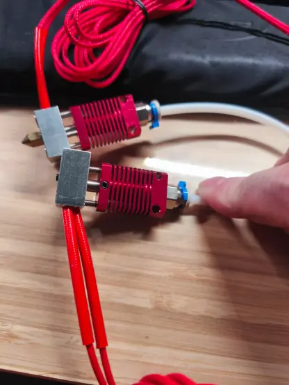 Zwei rote MK8 Hotends liegen auf einem Holzbrett. zwei rote "Kabel" raken aus dem HEizblock heraus (24V Heizung). Eines der Hotends wird durch einen Finger dazu gebracht ein schöneres Foto davon machen zu können.