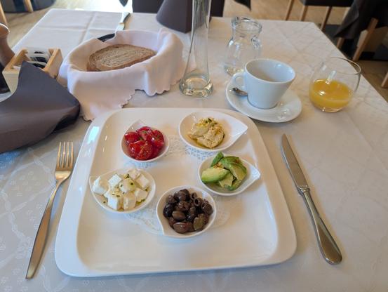 Frühstück "Mediterranae" im Restaurant, mit Kaffee, Orangensaft, Brot und einzelnen Schälchen mit Feta, Oliven, Tomaten, Avocado und Hummus.