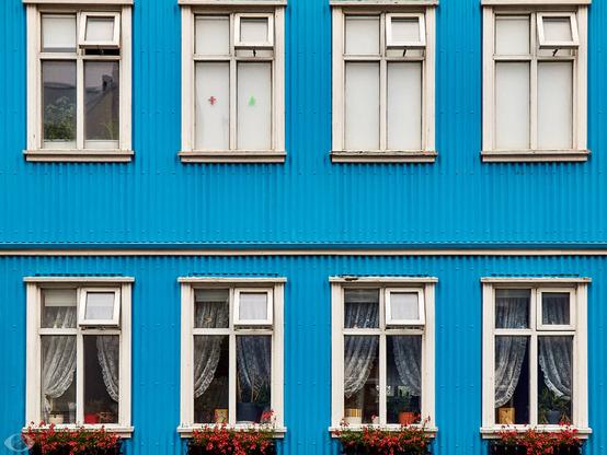 Blue wooden facade with eight windows in two rows of 4. The lower 4 have a flower box with red flowers beneath the balcony and a pair of open curtains behind each window. The upper windows lack that.