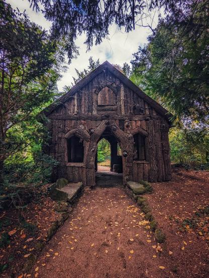 A charmingly rustic Borkenhäuschen (bark house) in Oranienbaum-Wörlitz, Germany, captured on a slightly overcast day. The small, wooden structure is fashioned from gnarled logs and branches, giving it a fairy-tale or witch-house aesthetic. A dirt path, scattered with autumn leaves, leads up to its central arched entrance. Two small, rustic benches made of stone and moss sit on either side of the entrance. The house is nestled amongst dense green trees and foliage, with dappled light filtering through the leaves above, enhancing its whimsical and secluded feel.