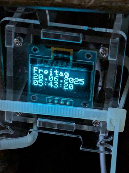 Ein mit Kabelbinder befestigtes ca. fünf cm würfelförmiges transparentes Plastik Gehäuse. Darin enthaltenen auf einer Platine ESP8266-01s und ein 0.96 OELD Display IIC. Als WLAN Netzwerk Uhr wird der Wochentag, das Datum und die Uhrzeit angezeigt.