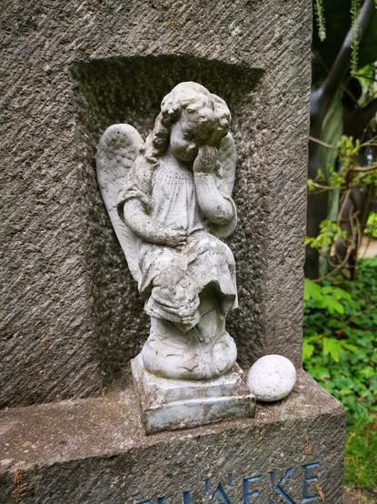 Ein steinerner, sitzender Engel mit lockigem Haar und Flügeln ist in eine Wandnische eingelassen. Der Engel stützt sein Kinn nachdenklich mit der linken Hand ab. Rechts neben der Engelsstatue liegt ein kleiner, glatter, weißer Stein. Im Hintergrund sind verschwommen grüne Pflanzen zu sehen, und der untere Teil der Wand, in der sich die Nische befindet, ist sichtbar.