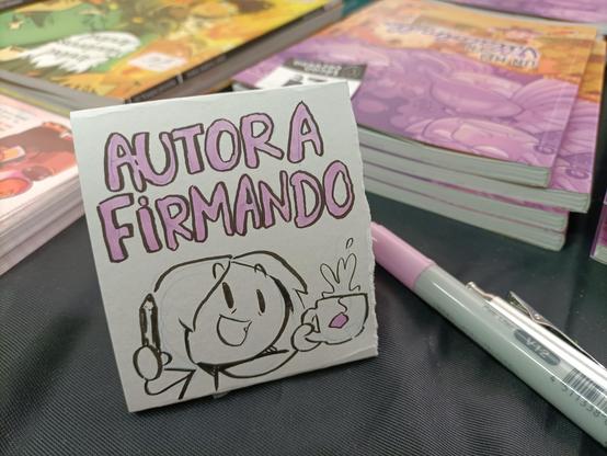 Foto de un cartel pequeño de papel sobre una mesa que pone "autora firmando" y un dibujo de Laurielle brindando con té