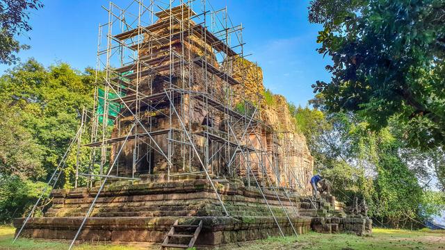 Photo shows a three-tower temple called Prasat Bat Chum in early Khmer architectural style. It is covered by scaffolds caused by a several yearlong restoration process. It is surrounded by tall, green forest and the platform for the towers stands on a green grass covered ground. It is a sunny morning with a visible clear blue sky.