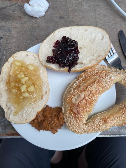 Ein Teller, Messer und Gabel, auf dem Teller zwei Brötchenhälften mit Marmelade, ein Happen Bohnencreme und ein Sesamteilchen