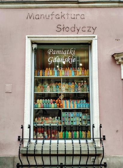 A white-framed, almost floor-to-ceiling window in a light brown and pink painted house wall. "Manufaktura Slodyczy" is written on the wall above the window and "Pamiatki Gdańskie" is written in white curved lettering at the top of the window pane. On the five shelves are mainly colourful miniature terraced houses made of clay or paper, modelled on Gdańsk town houses, and the small stone windowsill at the bottom is covered with an ornate cast-iron grille.
