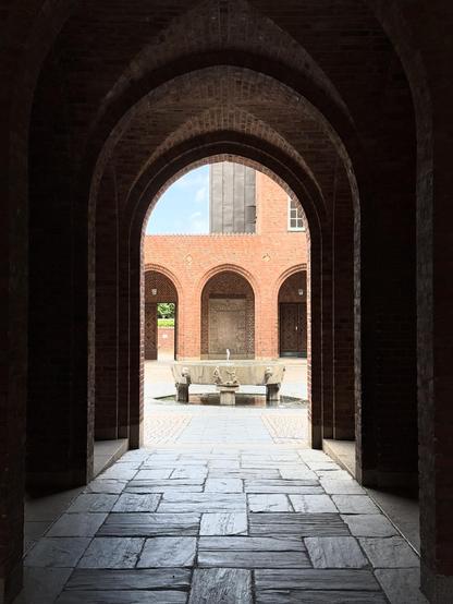 Looking through a brick archway and cloister towards a central fountain. The brick is red and without ornamentation.