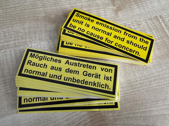 A stack of yellow caution labels displaying text in English and German, stating: „Smoke emission from the unit is normal and should be no cause for concern.“