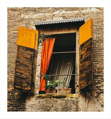 This photo is of a window set into a weathered brick wall.  The window is open with dark wooden shutters angled outward on either side. The top of the shutters are painted a yellow/ochre colour. There is a vibrant red curtain that hangs to the left hand side, partially obscuring the view inside. Inside there are sheer beige curtains visible. Two potted plants with white flowers sit on the windowsill. They have bright green leaves.