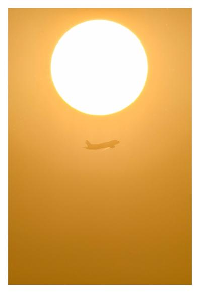 Foto im Hochformat. Teleaufnahme. Die leuchtende gelbe Sonne ist mittig oben im Bild. Der Himmel, der das restliche Bild ausfüllt ist gelblich orange. Direkt mittig unter der Sonne fliegt ein Passagierflugzeug, es steigt gerade von links nach rechts auf.