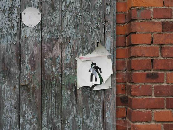 Worn wooden door with peeling paint, featuring a partially torn paper sign depicting a stylized figure holding an object. Adjacent to the door is a red brick wall with visible mortar lines. A small circular metal object is mounted on the door above the sign.