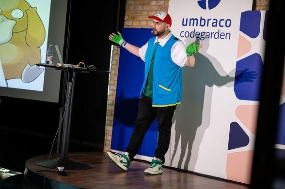 A speaker dressed as Ash Ketchum from Pokémon—complete with red cap, blue vest, and green gloves—presents on stage at Umbraco Codegarden. A large image of the Pokémon Psyduck is displayed on the screen beside him.